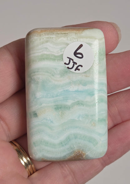A hand holding a polished rectangular stone with rounded corners, showing a white and pale green marbled pattern. A small white sticker with the number 6 is attached to one side.