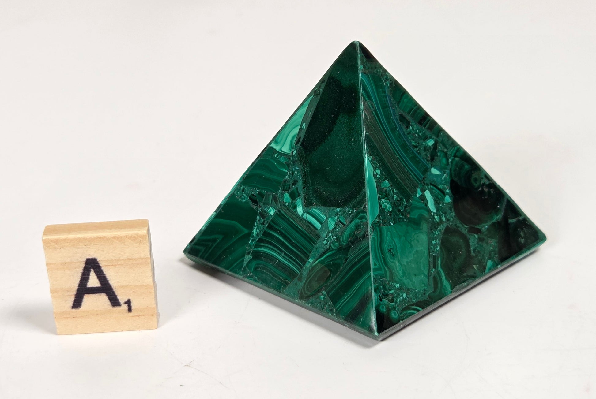 A polished green malachite crystal pyramid with natural stone banding, shown next to a small wooden letter tile spelling 'A1' on a plain white background.