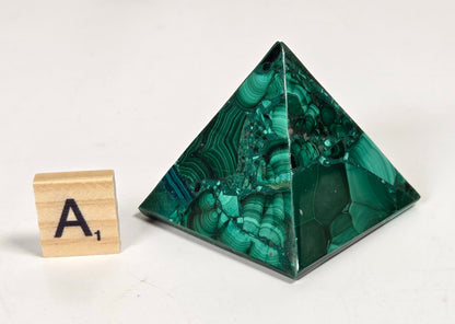 A polished green malachite crystal pyramid with natural banded patterns, shown next to a wooden Scrabble tile with the letter 'A' on a white background.