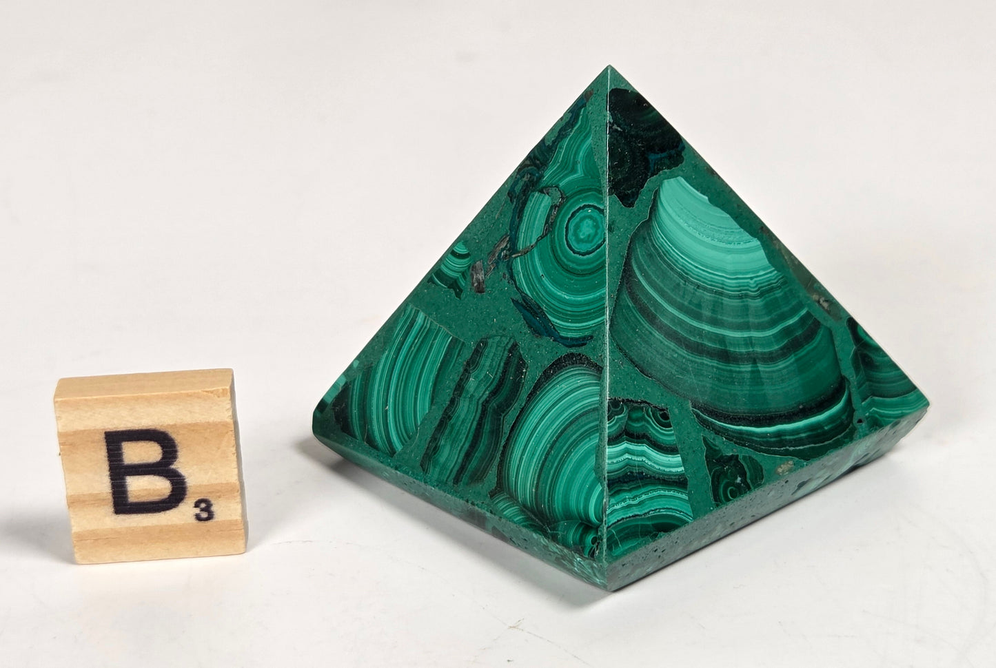 A polished green malachite crystal pyramid with natural banded patterns, shown next to a wooden Scrabble tile with the letter 'B' on a white background.