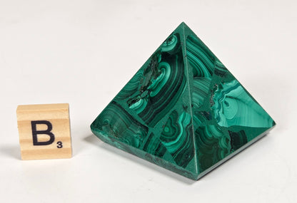 A polished green malachite crystal pyramid with natural swirling patterns, shown next to a small wooden Scrabble tile displaying the letter 'B' on a clean white background.