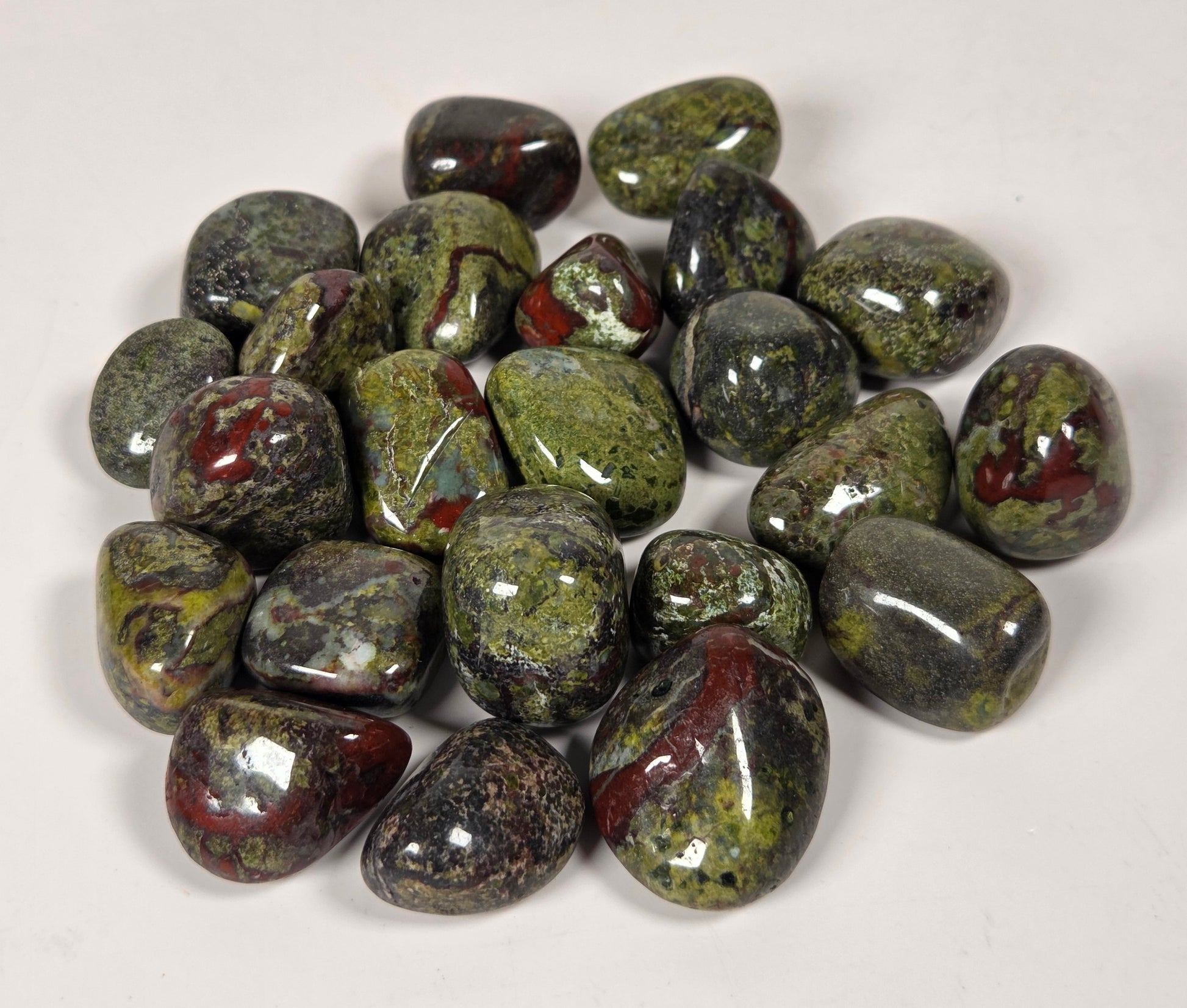 A pile of polished tumbled gemstones in green, black, and reddish-brown marbled patterns, photographed on a plain off-white background.