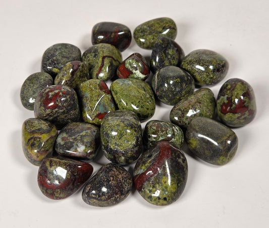 A pile of polished tumbled gemstones in green, black, and reddish-brown marbled patterns, photographed on a plain off-white background.