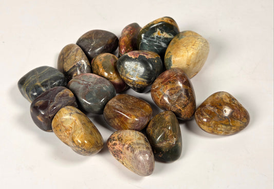 A pile of polished tumbled stones in earthy brown, beige, gray, and green tones, arranged on a plain off-white background.