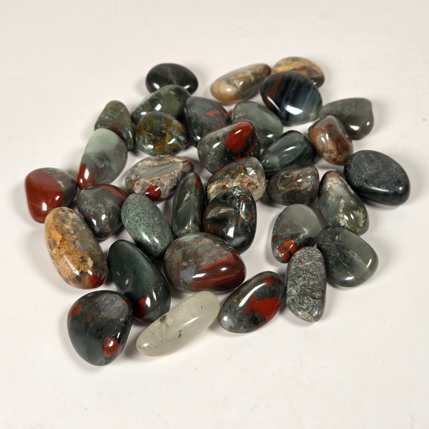 A pile of polished tumbled gemstones in various earthy colors including gray, green, black, brown, and red, arranged on a plain off-white background.
