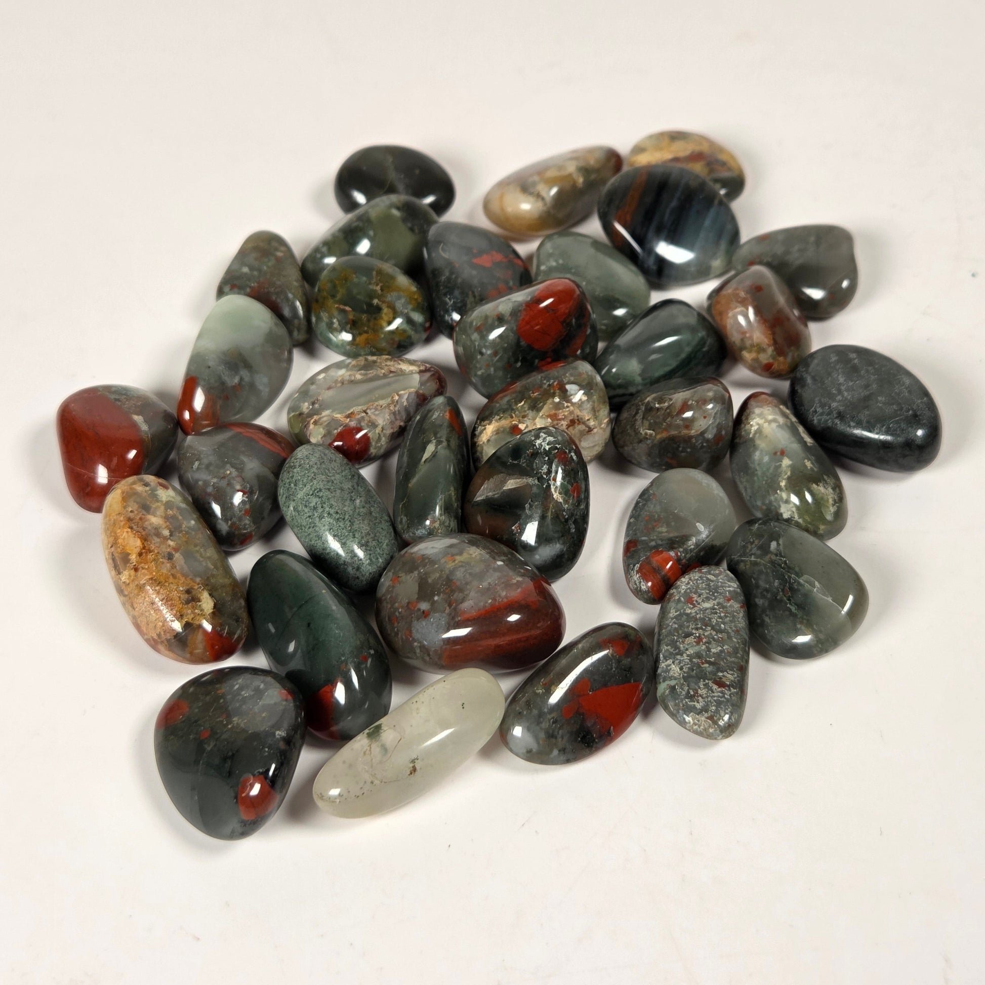 A pile of polished tumbled gemstones in various earthy colors including gray, green, black, brown, and red, arranged on a plain off-white background.