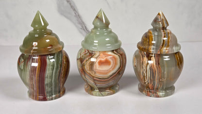 Three small decorative lidded jars made from polished agate stone, featuring swirling patterns of green, brown, beige, and reddish tones, arranged side by side on a white surface with a white and gray marble background.