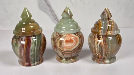 Three small decorative lidded jars made from polished agate stone, featuring swirling patterns of green, brown, beige, and reddish tones, arranged side by side on a white surface with a white and gray marble background.