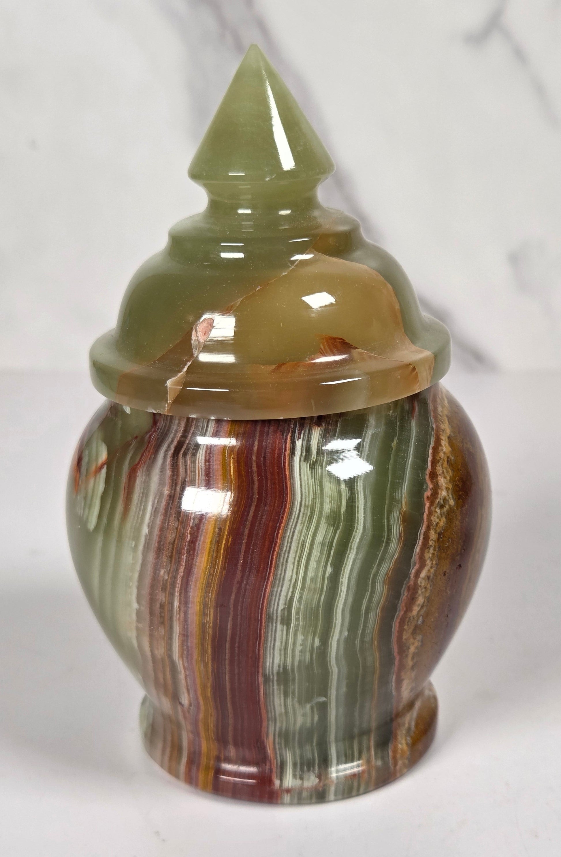 small decorative lidded jar made from polished agate stone, featuring swirling patterns of green, brown, beige, and reddish tones, on a white surface with a white and gray marble background.