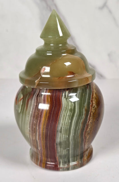 small decorative lidded jar made from polished agate stone, featuring swirling patterns of green, brown, beige, and reddish tones, on a white surface with a white and gray marble background.