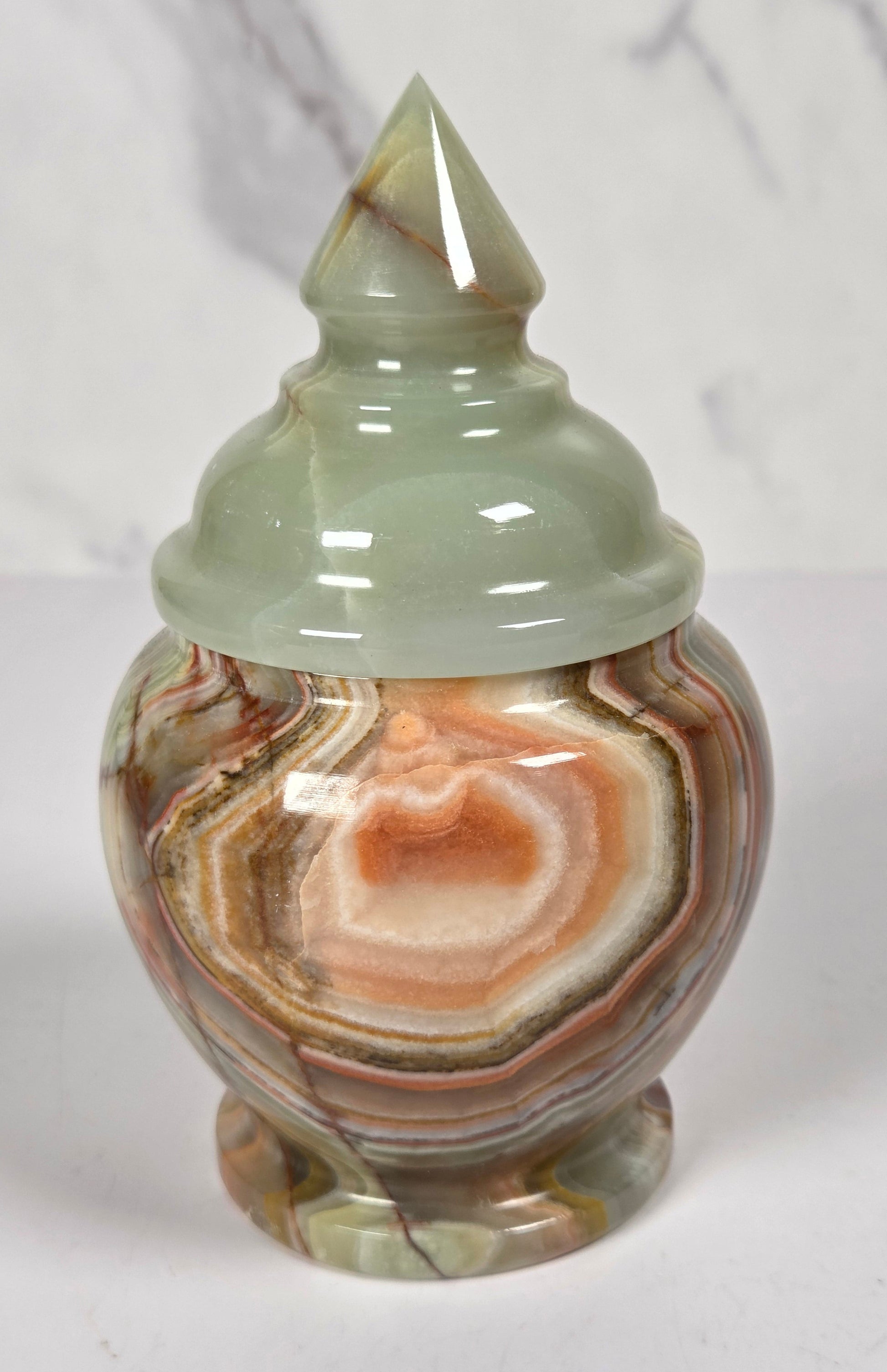 small decorative lidded jar made from polished agate stone, featuring swirling patterns of green, brown, beige, and reddish tones, on a white surface with a white and gray marble background.