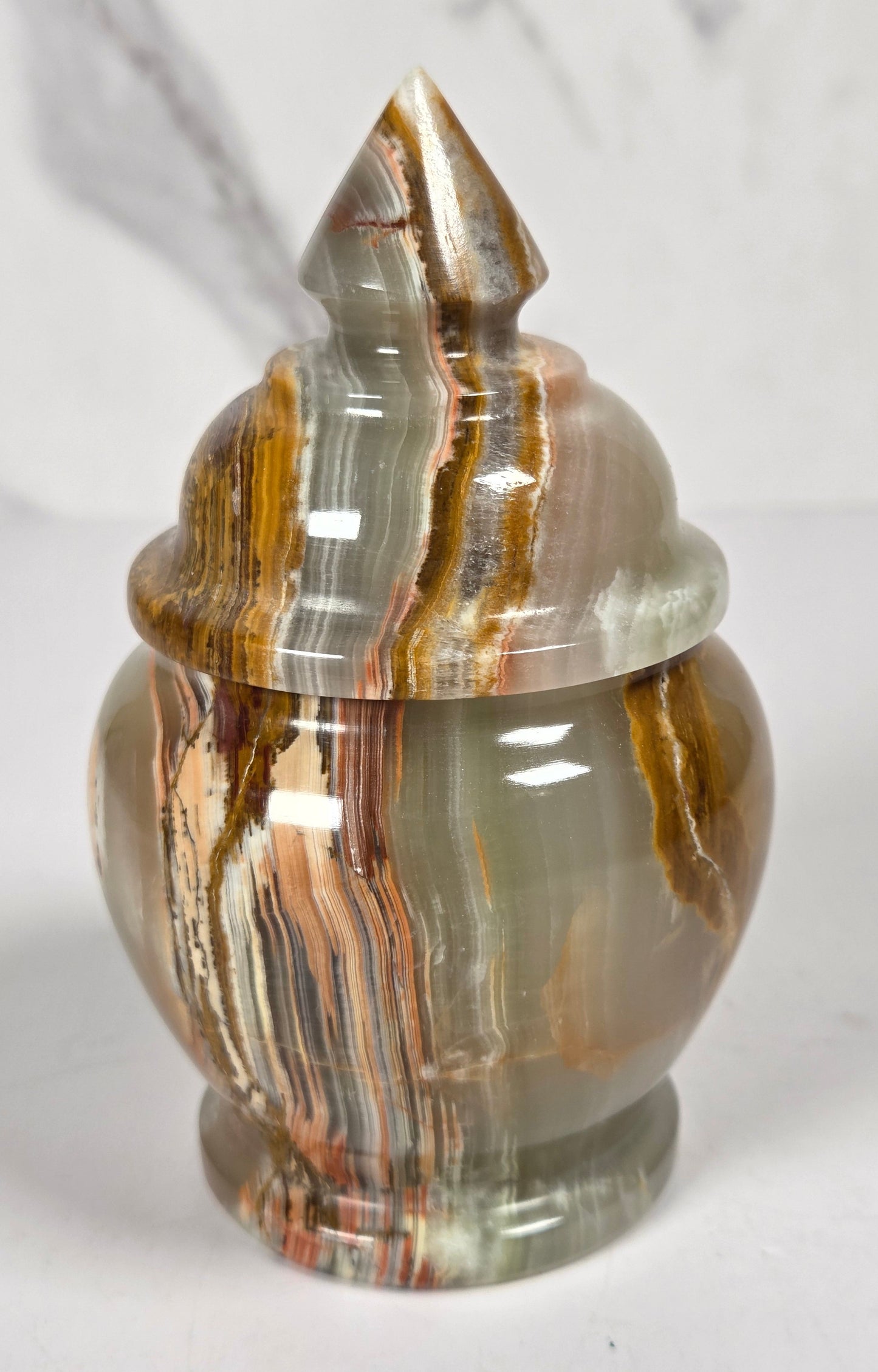small decorative lidded jar made from polished agate stone, featuring swirling patterns of green, brown, beige, and reddish tones, on a white surface with a white and gray marble background.