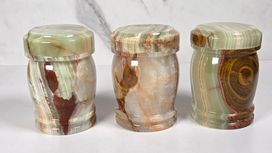 small decorative lidded jar made from polished agate stone, featuring swirling patterns of green, brown, beige, and reddish tones, on a white surface with a white and gray marble background.