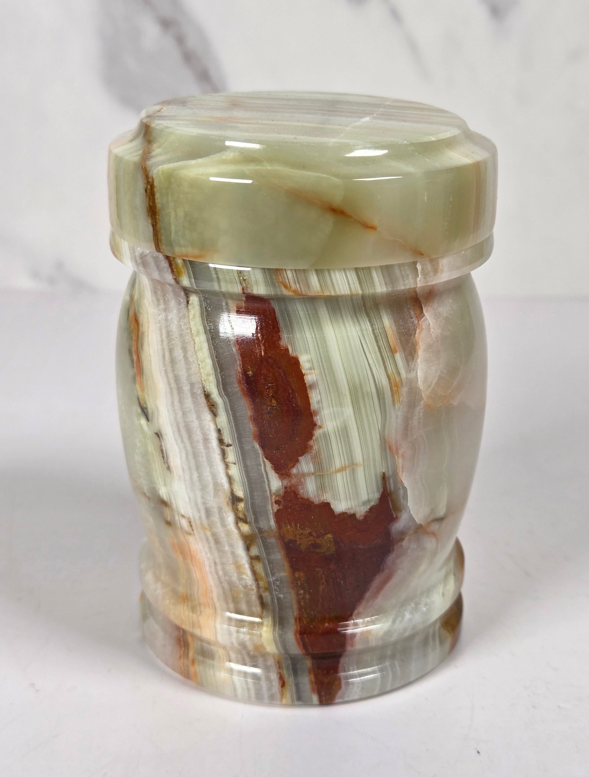 small decorative lidded jar made from polished agate stone, featuring swirling patterns of green, brown, beige, and reddish tones, on a white surface with a white and gray marble background.