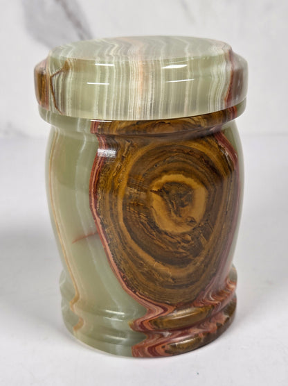 small decorative lidded jar made from polished agate stone, featuring swirling patterns of green, brown, beige, and reddish tones, on a white surface with a white and gray marble background.