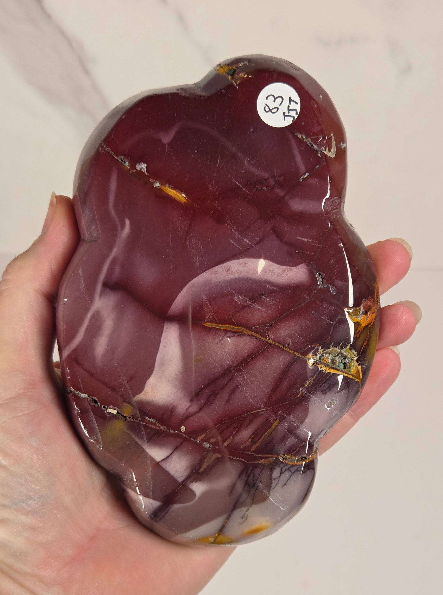 A hand holding a polished, organic-shaped piece of dark reddish-brown stone or crystal with natural marbling and small gold inclusions, against a light off-white marble background.