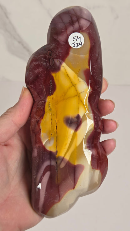 A hand holding a polished natural stone carving with marbled dark red, yellow, and cream sections, photographed against a light indoor wall.