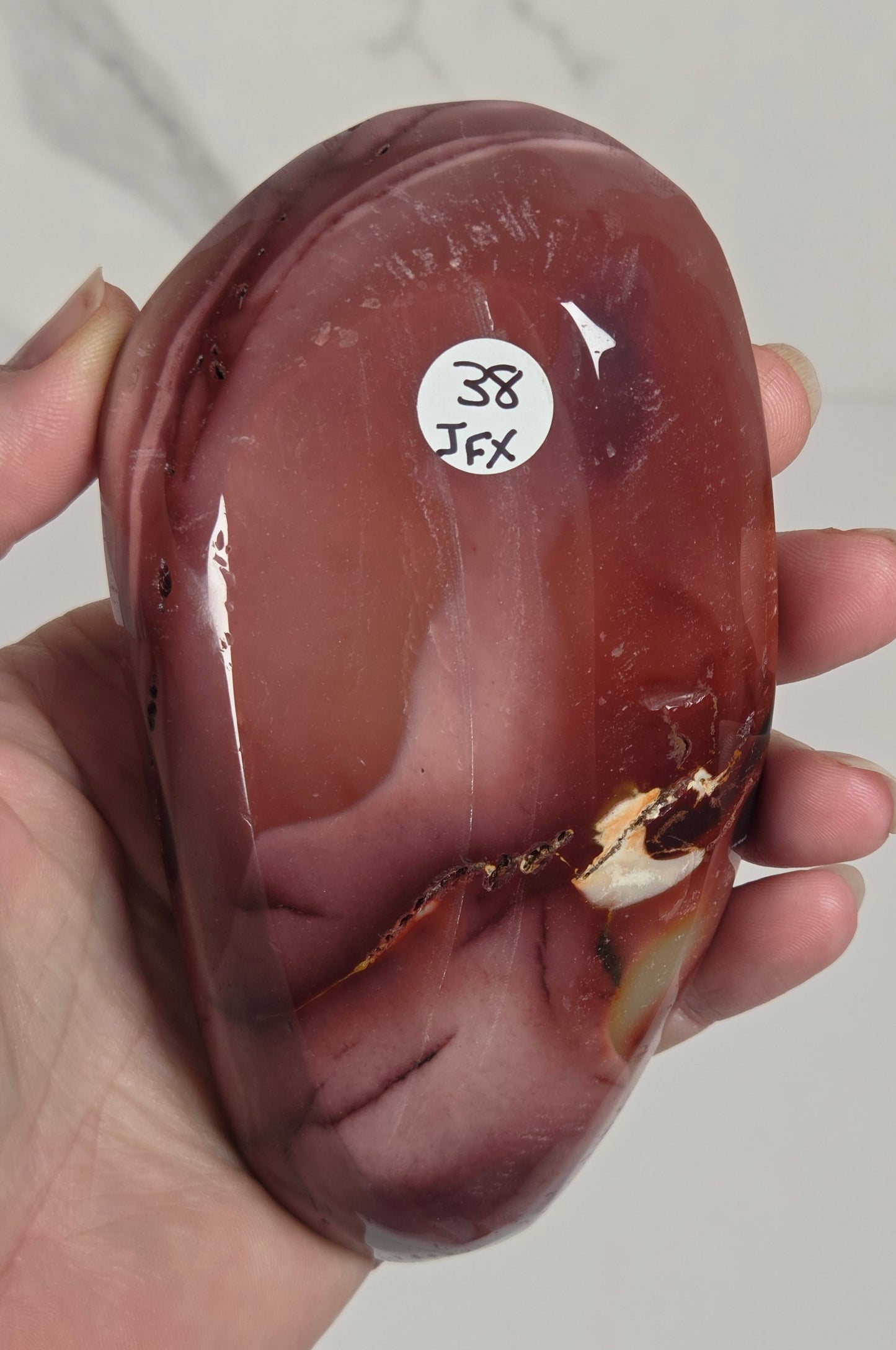 A hand holding a polished reddish-brown stone or agate slab with natural marbling and a small white sticker label on the surface, photographed against a light neutral background.