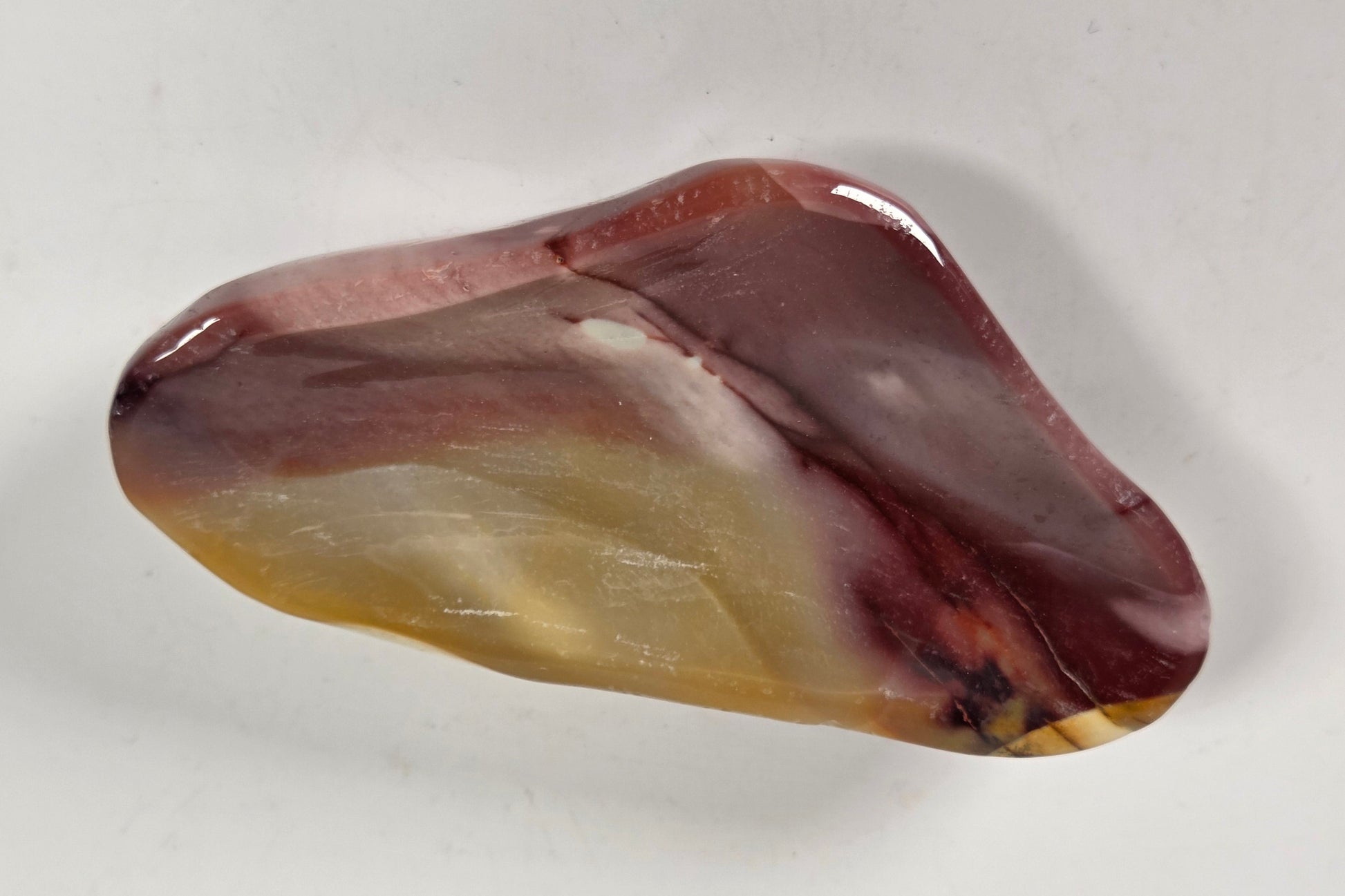 A polished, irregular-shaped stone or crystal with marbled patterns of reddish-brown, amber yellow, and white, displayed on a plain light gray background.