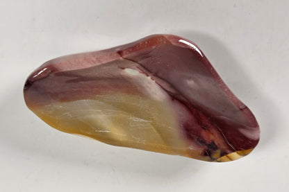 A polished, irregular-shaped stone or crystal with marbled patterns of reddish-brown, amber yellow, and white, displayed on a plain light gray background.