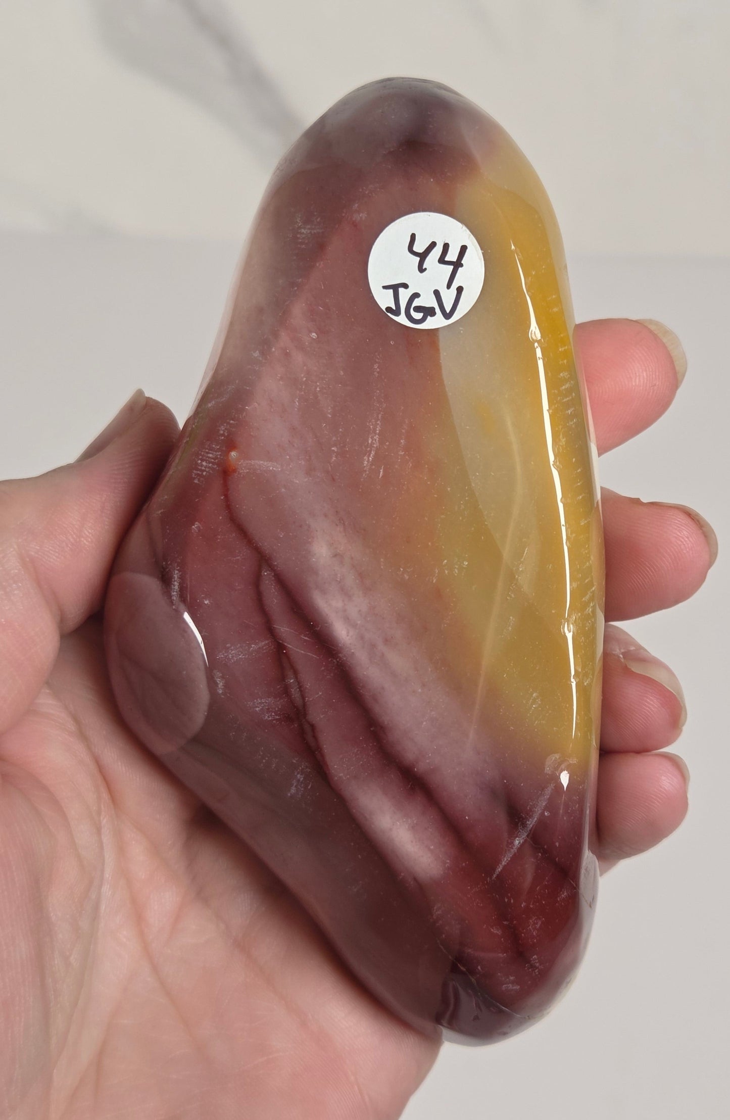 A hand holding a polished, smooth stone with a color gradient from yellow-gold at the top to deep reddish-brown at the bottom, featuring a small white sticker with handwritten text on the left side.