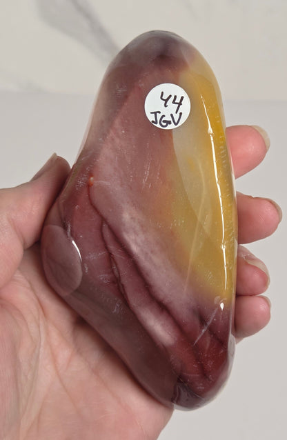 A hand holding a polished, smooth stone with a color gradient from yellow-gold at the top to deep reddish-brown at the bottom, featuring a small white sticker with handwritten text on the left side.