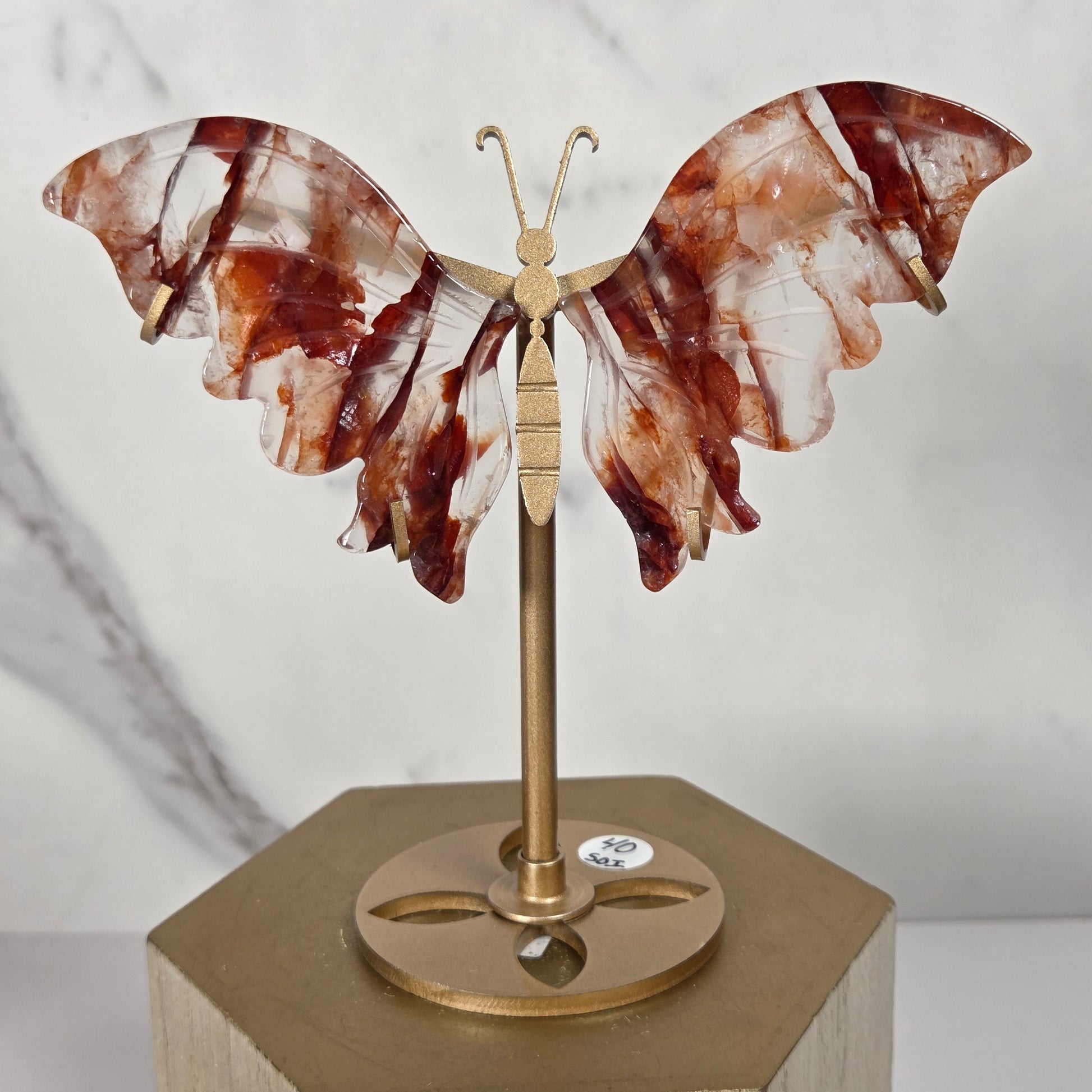 A decorative wall-mounted butterfly sculpture with translucent brown and white marbled wings on a brass rod, attached to a hexagonal light wood wall bracket against a white marble-patterned background.