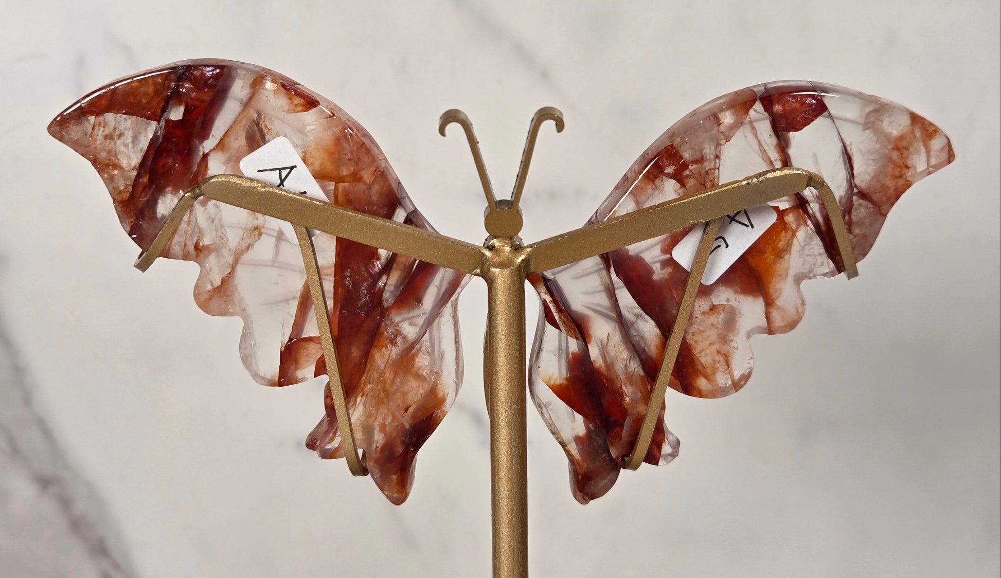 A butterfly-shaped carved piece with translucent brown and white marbled wings mounted on a gold-tone metal stand, photographed against a light marble-patterned background.