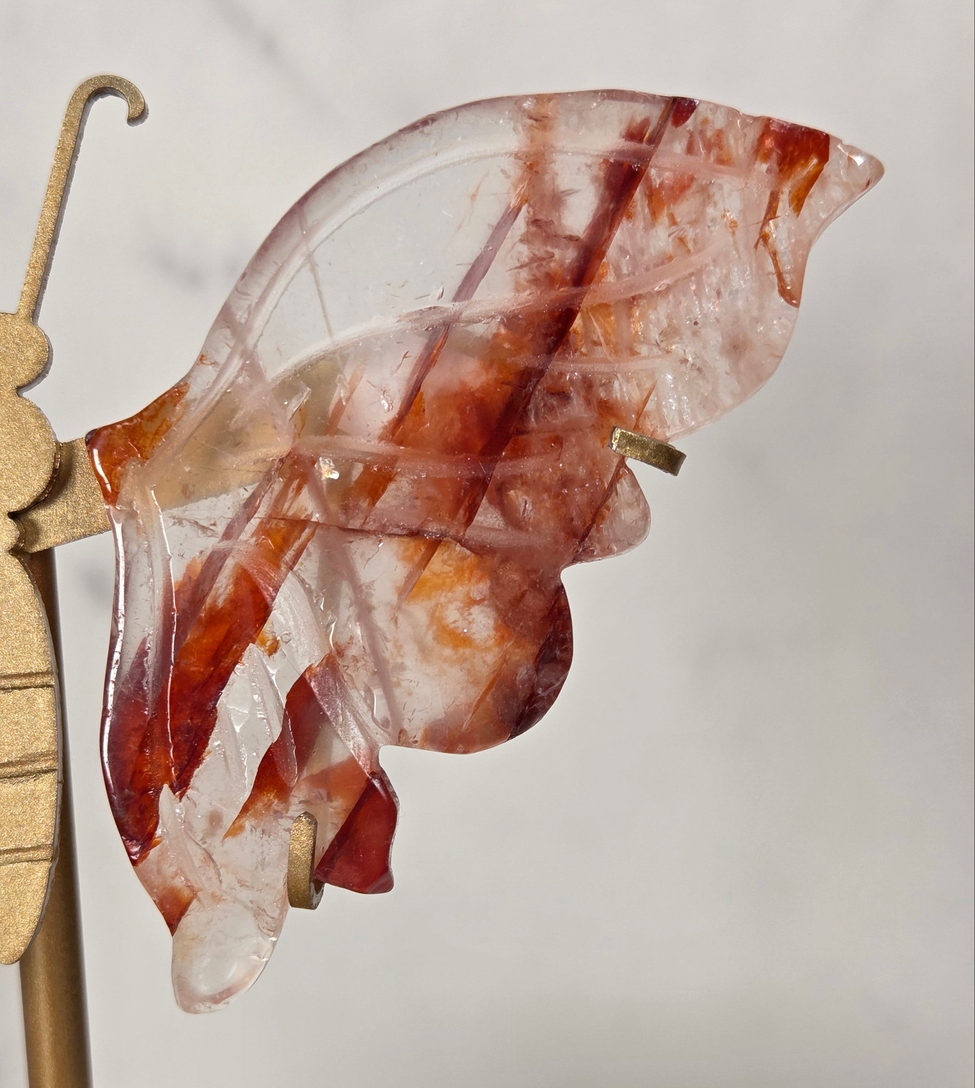 A close-up of a decorative butterfly-shaped wall hanging made from translucent reddish-brown stone wings attached to a brass rod and finial, shown against a light neutral background.