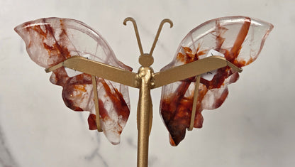 A butterfly-shaped jewelry stand or display holder made of gold-toned metal with translucent wings featuring brown and white marbled patterns, set against a light marble background.