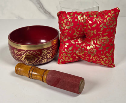 A Tibetan singing bowl in red with a floral pattern on the rim with a cushion and a striker.