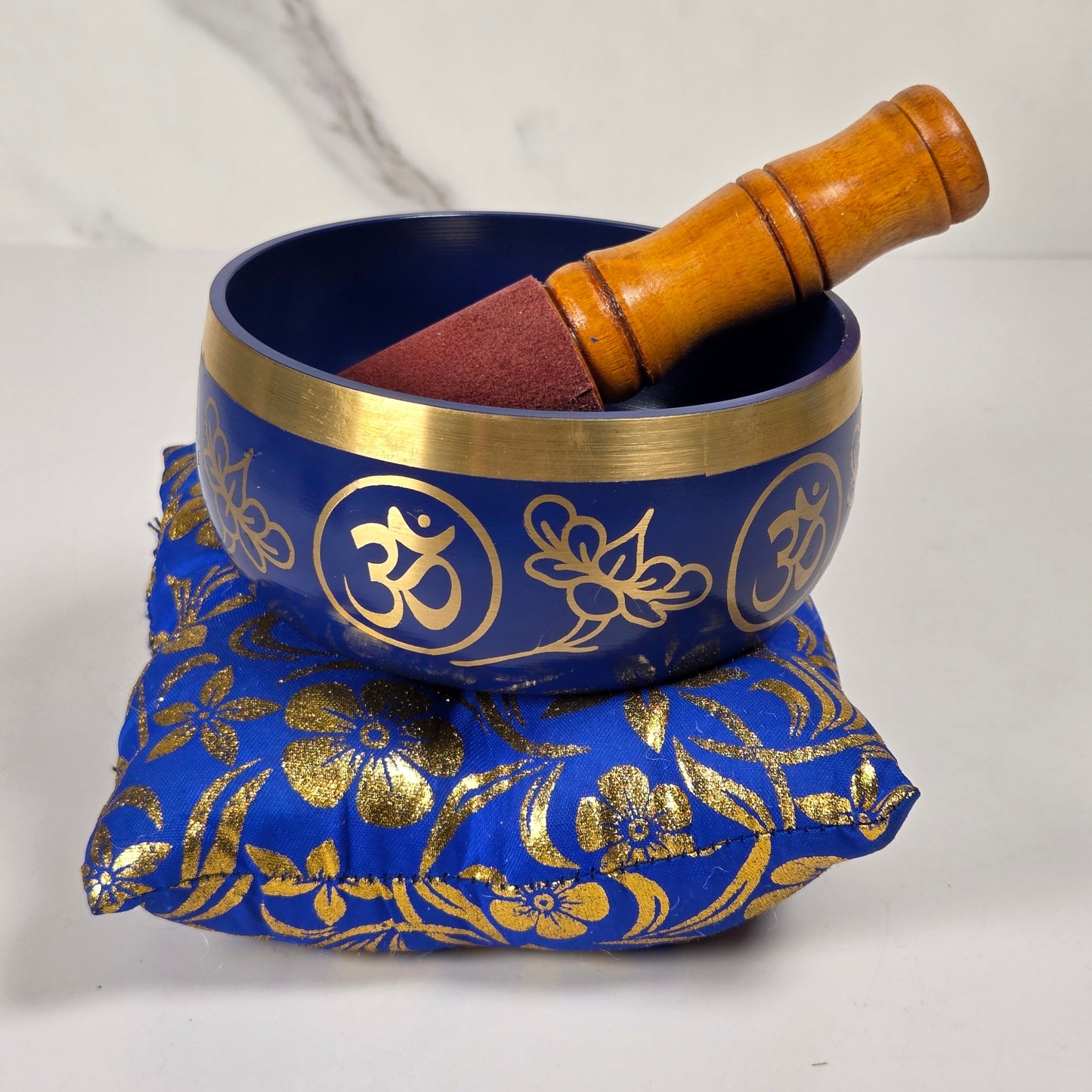 A blue and gold brass singing bowl with a wooden mallet and a blue fabric cover decorated with gold floral patterns, shown against a white marble background.