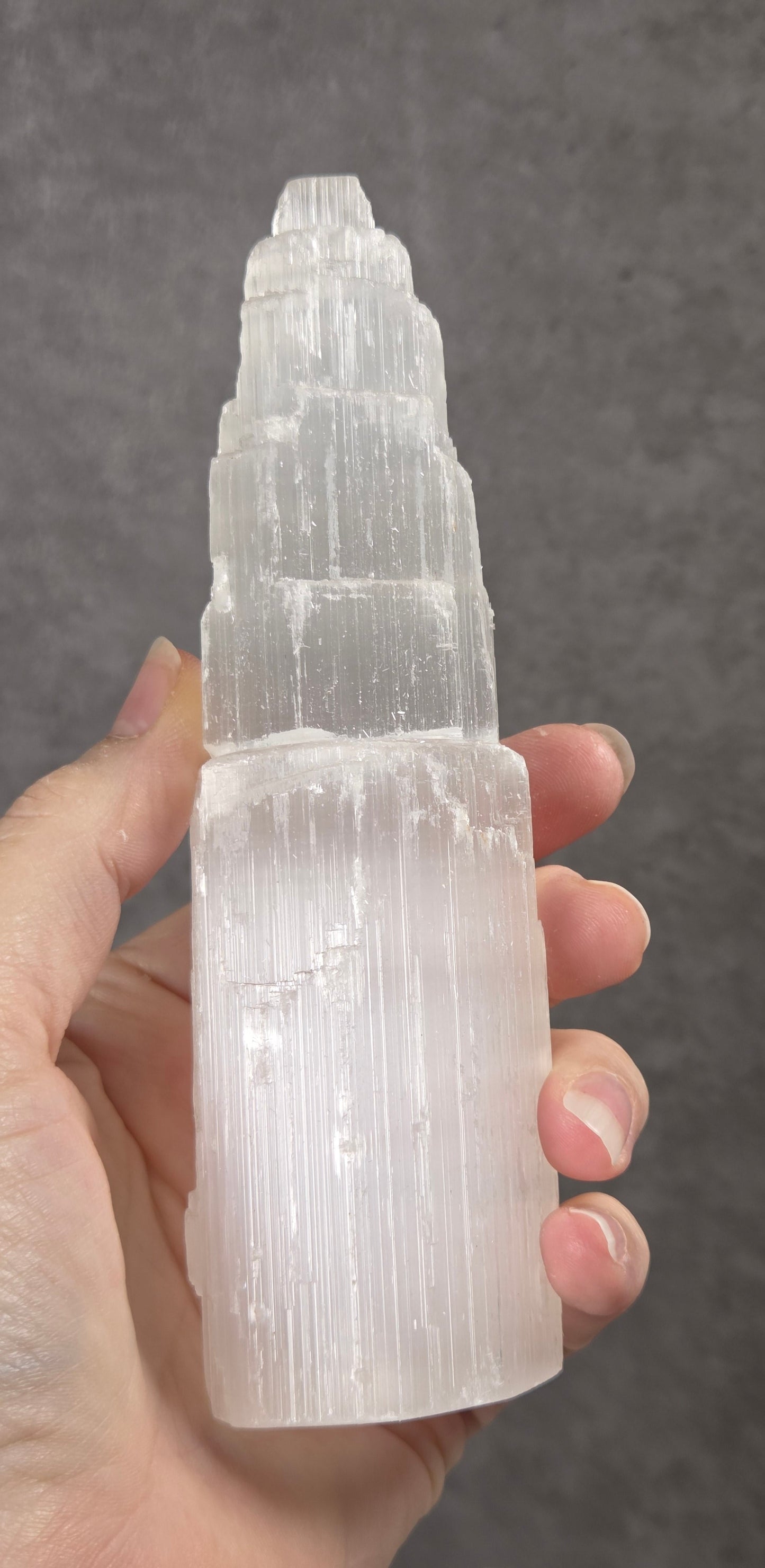 A hand holding a tall, tiered clear crystal tower, likely a selenite or quartz cluster, against a dark textured gray background.