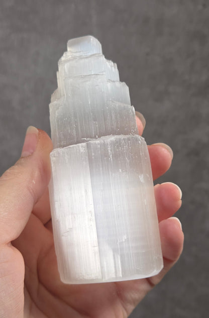 A hand holding a white selenite tower crystal cluster against a textured gray background.