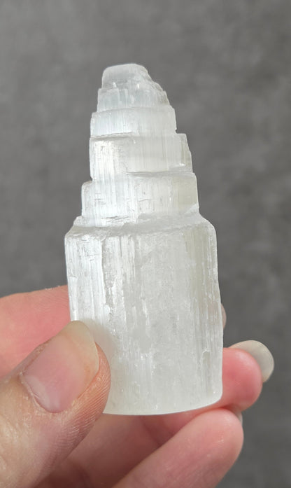 A hand holding a small, tapered white crystal cluster with a pointed top, photographed against a textured gray background.