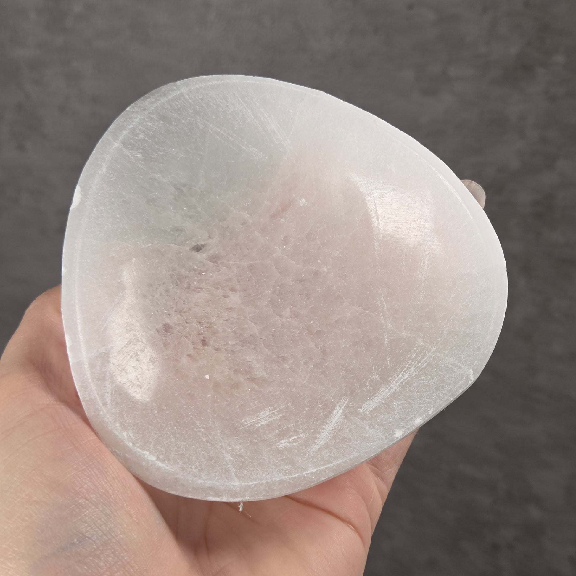 A hand holding a translucent white selenite bowl against a dark gray textured background.