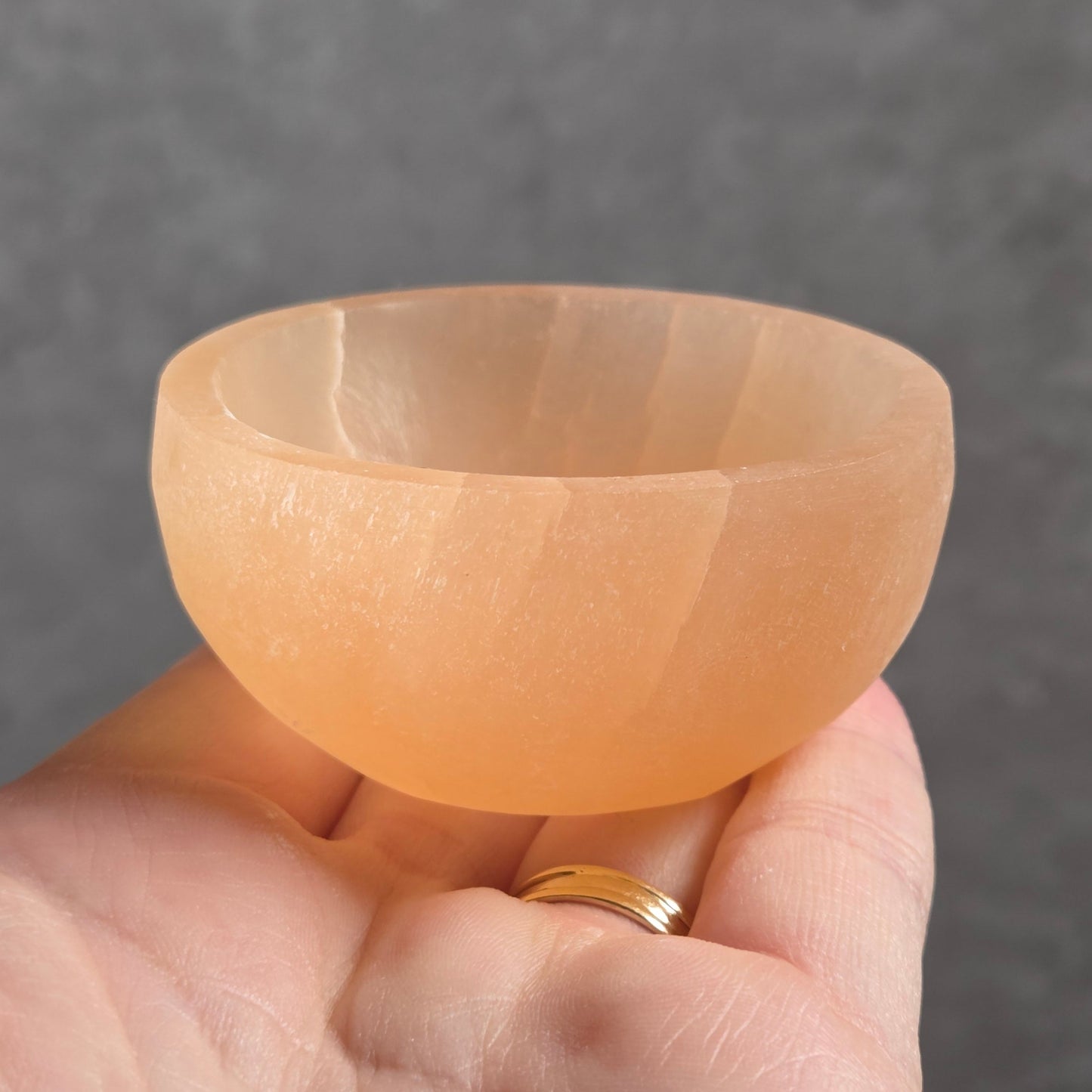 A hand holding a small, translucent peach-colored selenite bowl against a gray textured background.