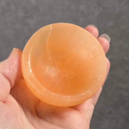A hand holding a small, translucent peach-colored selenite bowl against a gray textured background.