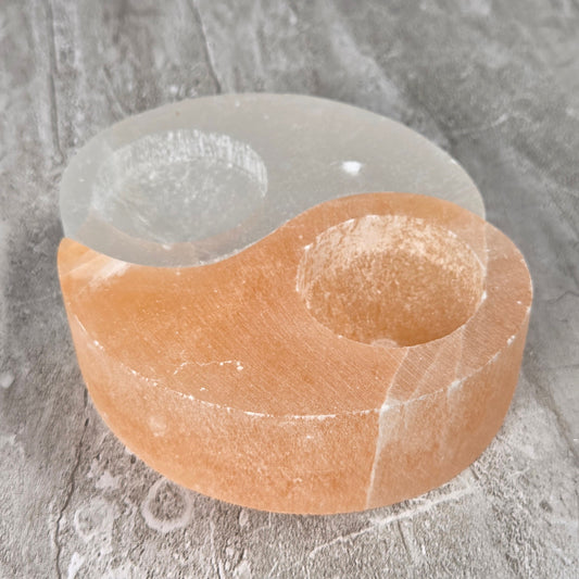 A two-compartment candle holder made from selenite and peach selenite, resting on a gray textured surface.