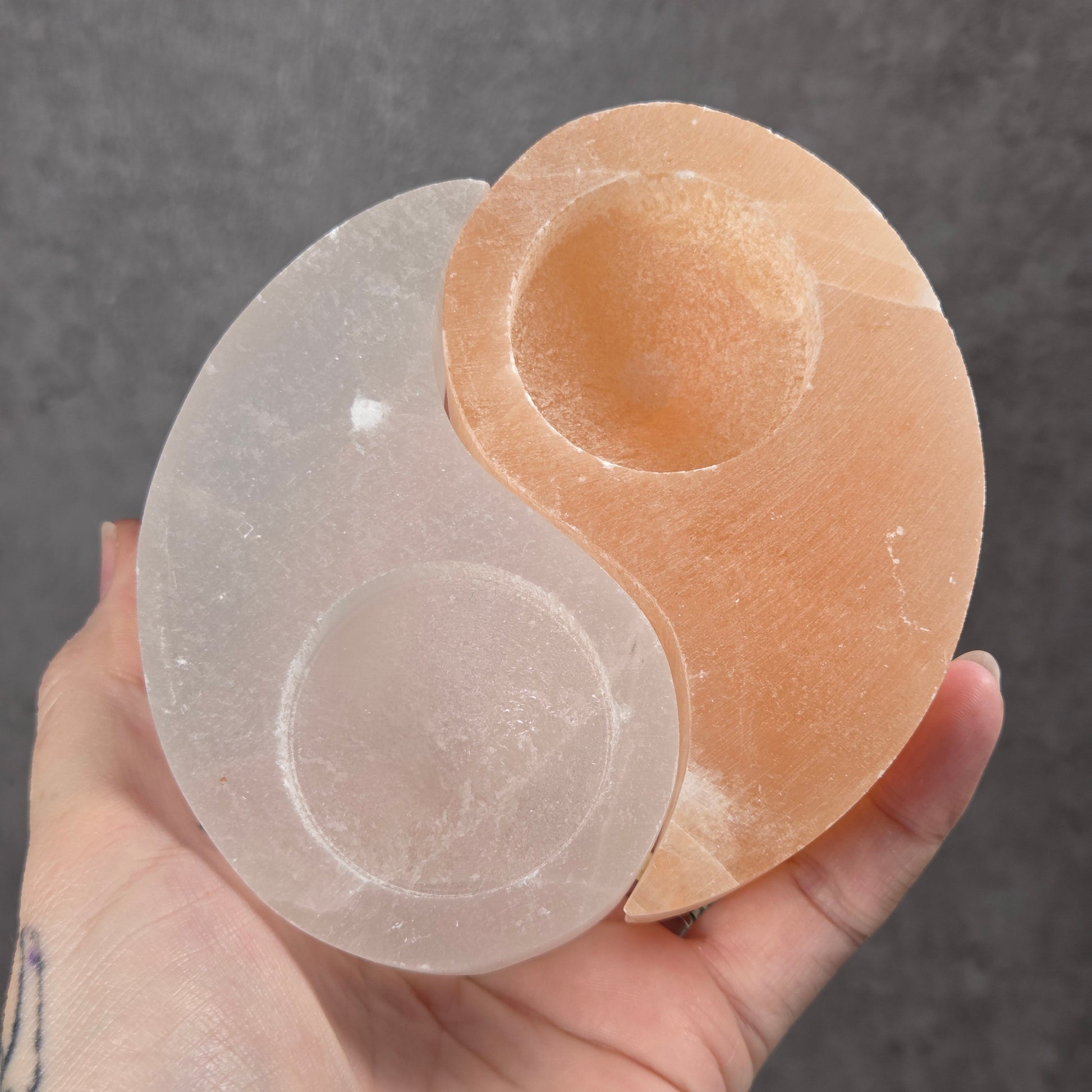 A hand holding a dual-toned salt crystal cup set shaped like a yin-yang, featuring one peach-colored cup and one milky white cup, against a textured gray background.