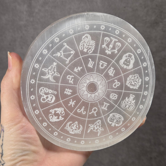 A hand holding a circular clear acrylic zodiac wheel with white engraved astrological symbols and text, set against a dark gray textured background.