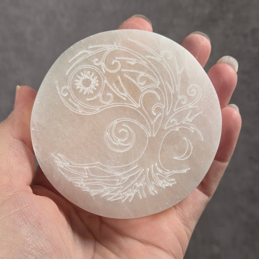 A hand holding a round, frosted white stone medallion engraved with a detailed white floral and scroll pattern, set against a textured gray background.