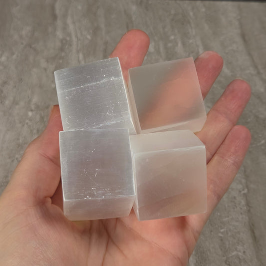 Four translucent white rectangular blocks, likely selenite crystals, held in a person's hand against a gray stone tile background.