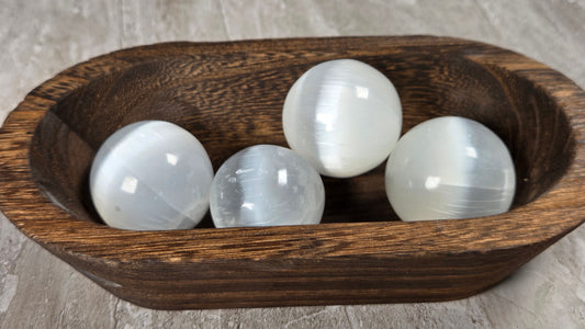 Four white selenite gemstone spheres resting in a dark brown wooden bowl on a textured gray stone surface.