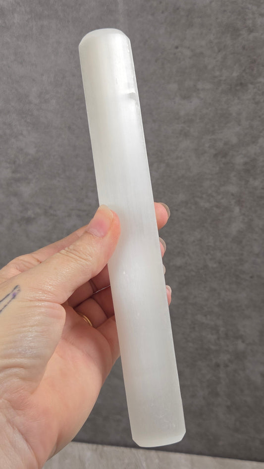 A hand holding a frosted, translucent white cylindrical selenite rod against a gray textured background.
