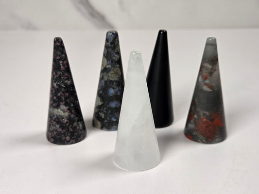 Five pointed crystal cone pendants in various colors and patterns, including black, gray, white, and marbled stone, arranged on a light surface.