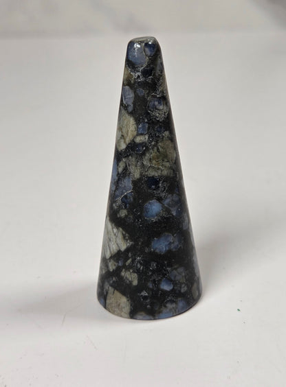 A natural polished blue sapphire cone-shaped cabochon, standing upright on a white surface with a blurred gray background.