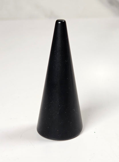 A small black cone-shaped object, possibly a candle holder or decorative object, standing on a white surface with a blurred gray background.