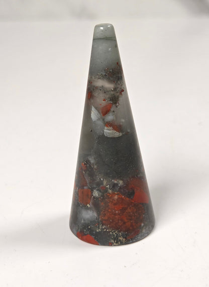 A small pointed cone-shaped stone or crystal with a gray translucent body and embedded red, white, and black inclusions, standing on a plain white surface.