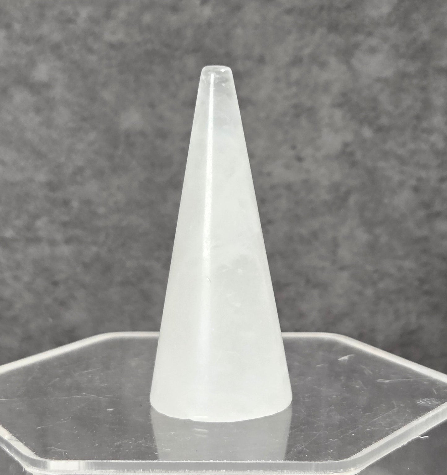 A frosted white cone-shaped object displayed upright on a clear hexagonal acrylic stand against a textured gray background.